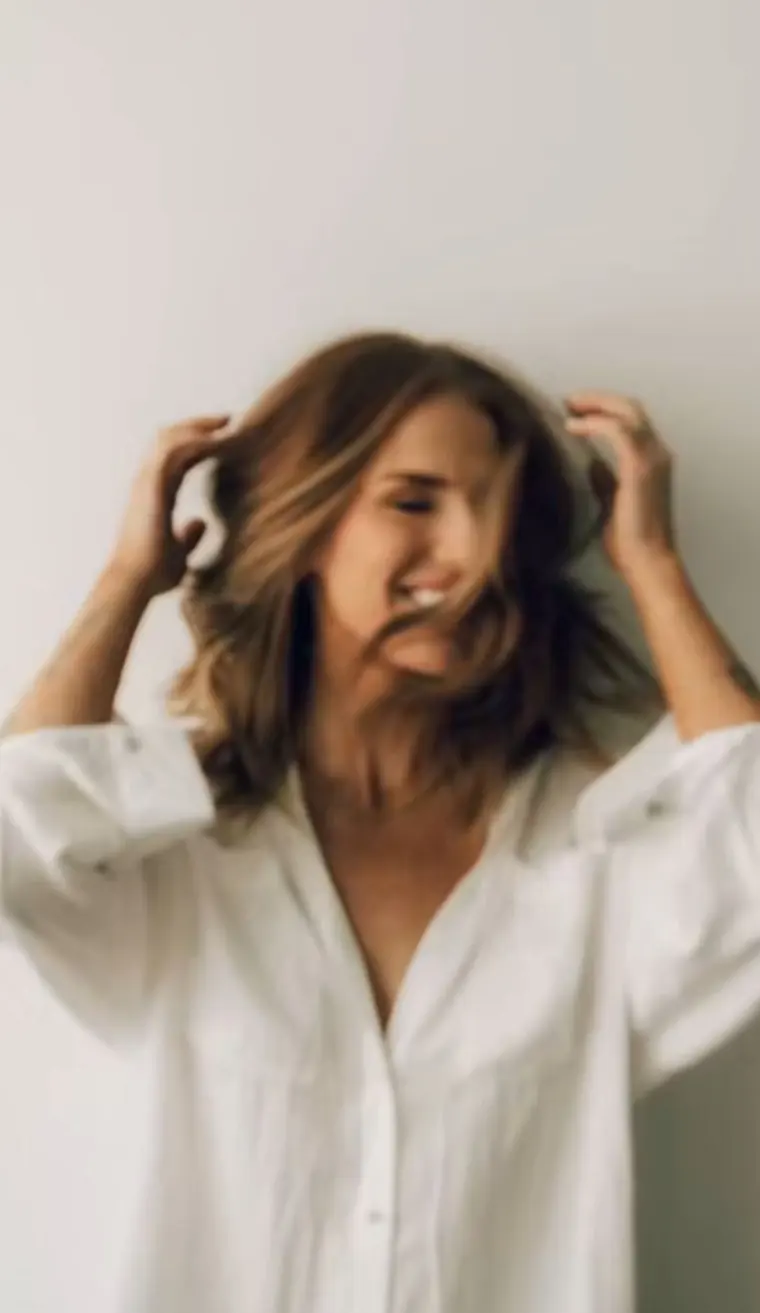 A woman standing against a white wall with her hands in her hair, laughing